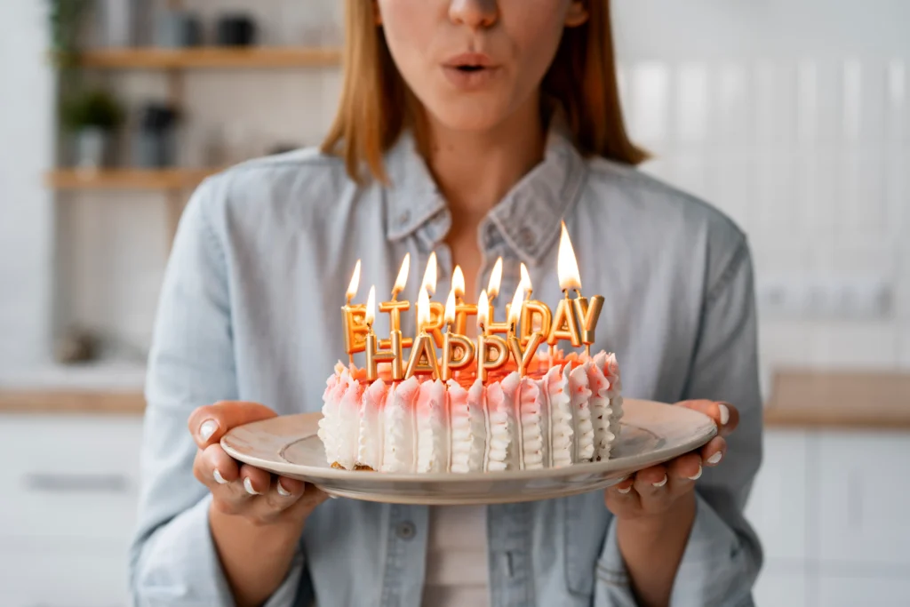 Mujer sosteniendo un pastel con velas encendidas que dicen HAPPY BIRTHDAY y soplando para apagarlas